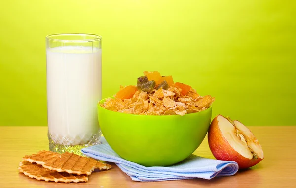 Смачні кукурудзяні пластівці в зеленій мисці, яблуко і склянка молока на дерев'яному столі на зеленому фоні — стокове фото