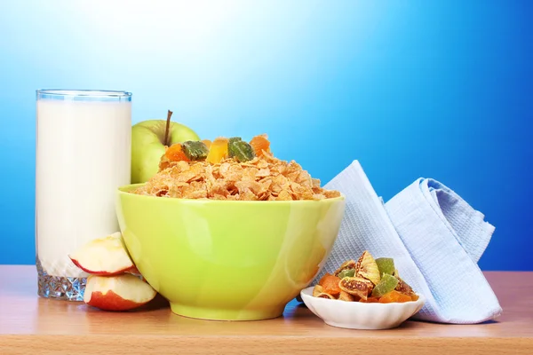 在绿色碗、 苹果和一杯牛奶在蓝色背景上的木桌上的美味玉米片 — 图库照片