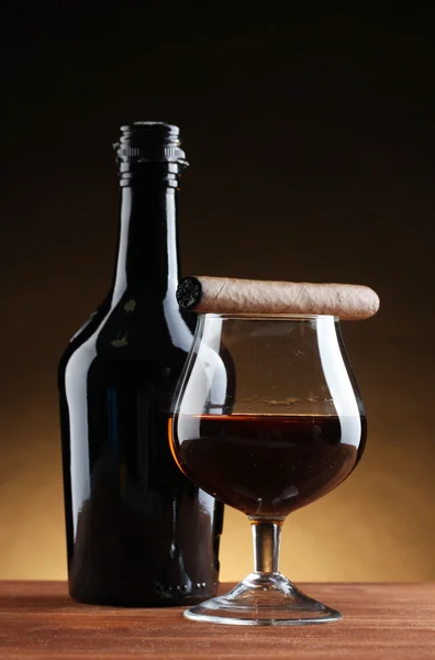 Botol dan gelas brendi dan cerutu di atas meja kayu dengan latar belakang coklat — Stok Foto