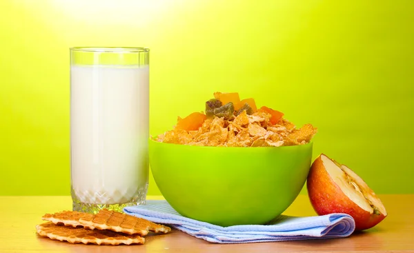 Tasty cornflakes in green bowl, apple and glass of milk on wooden table on green background — Stock Photo, Image