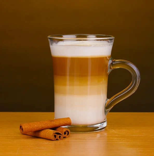 Fragrante cappuccino cappuccino cappuccino in tazza di vetro e cannella su tavolo di legno su fondo marrone — Foto Stock