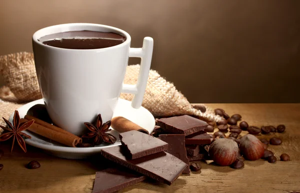 Tasse heiße Schokolade, Zimtstangen, Nüsse und Schokolade auf Holztisch auf braunem Hintergrund — Stockfoto