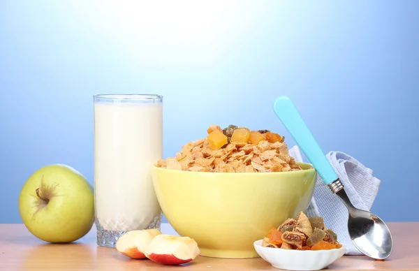 Tasty cornflakes in green bowl, apples and glass of milk on wooden table on blue background — Stock Photo, Image