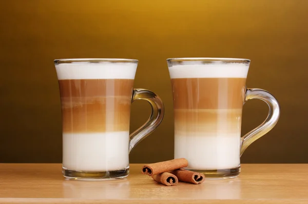 Pachnące? Unduli latte kubki szklane i cynamon na drewnianym stole na brązowym tle — Zdjęcie stockowe