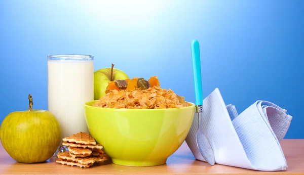 Smaczne płatki kukurydziane w zielonej miski, jabłka i szklankę mleka na drewnianym stole na niebieskim tle — Zdjęcie stockowe