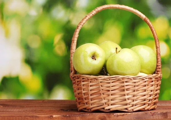 Saftigt gröna äpplen i korgen på träbord på grön bakgrund — Stockfoto