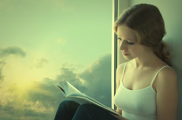 Beautiful young woman reading a book while sitting at a window i — Stock Photo, Image