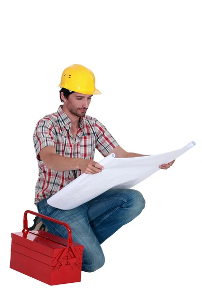 Builder reading plans whilst kneeling by tool-box — Stock Photo, Image