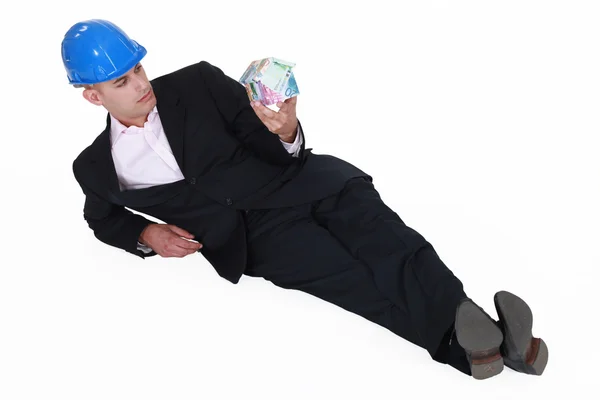 Engineer lazily lying on the floor and staring at a house made of money — Stock Photo, Image