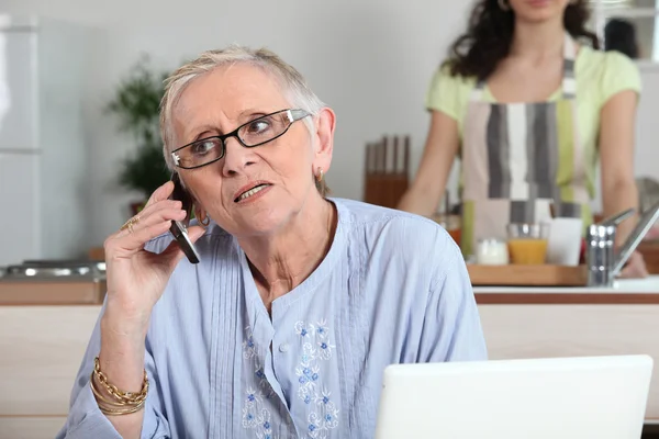 Ältere Frau telefoniert, während junge Helferin in der Küche arbeitet — Stockfoto