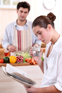 Young woman reading a newspaper while her boyfriend prepares lunch clipart