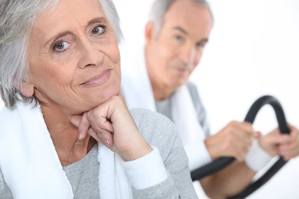 Elderly lady posing in fitness center with husband in background on exercise bike — Stock Photo, Image