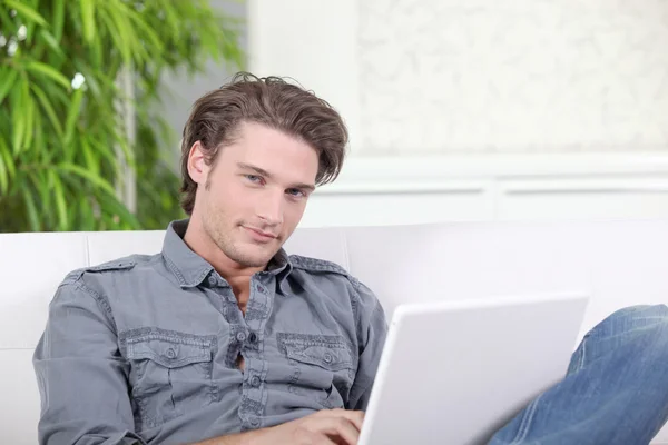 Bir dizüstü bilgisayar ile bir kanepede oturan genç adam — Stok fotoğraf