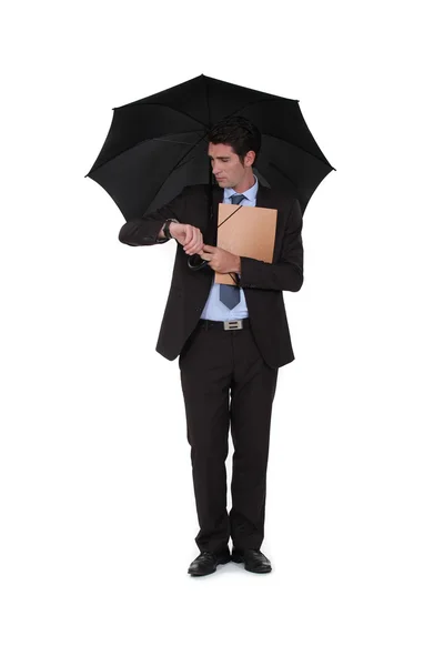 Ein Geschäftsmann unter einem Regenschirm, der auf seine Uhr schaut. — Stockfoto