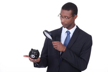 African man looking at a piggy bank through a magnifying glass clipart