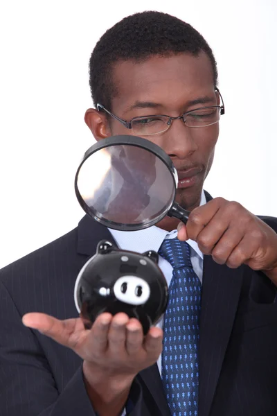 Zwarte goed geklede man kijken een spaarvarken met een vergrootglas — Stockfoto