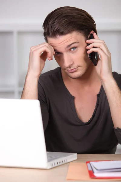 Young man in front of computer on the phone — Stock Photo, Image