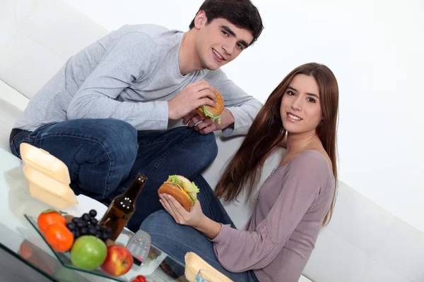 Jovem casal comer hambúrgueres no sofá — Fotografia de Stock
