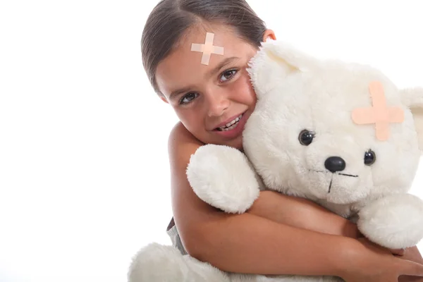 Jong meisje en haar teddybeer, beide dragen bijpassende pleisters op hun voorhoofd — Stockfoto