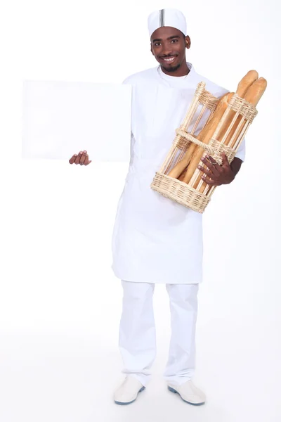 Panadero joven con una cesta de baguettes y una tabla en blanco para su mensaje — Foto de Stock