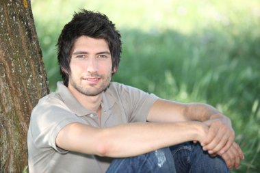 In the grass, a young man seated against a tree clipart