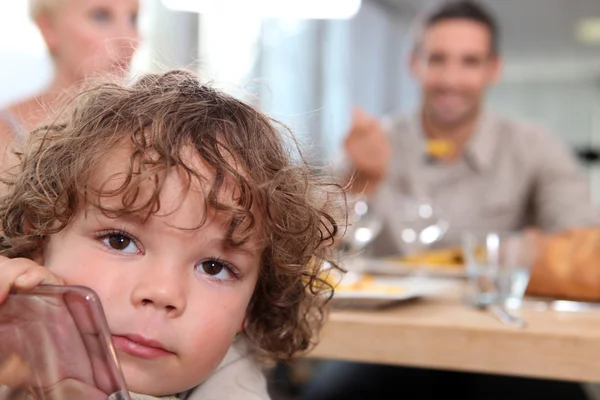 Mały chłopiec siedzi przy stole z rodzicami w tle — Zdjęcie stockowe