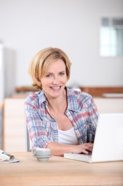 Woman sat in kitchen typing on laptop computer clipart