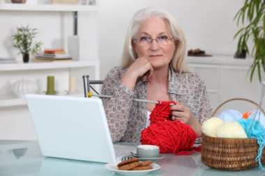 Elderly woman knitting by her laptop clipart