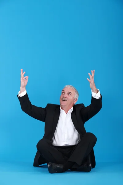 Mature man sitting cross-legged with raised arms against blue background — Stock Photo, Image
