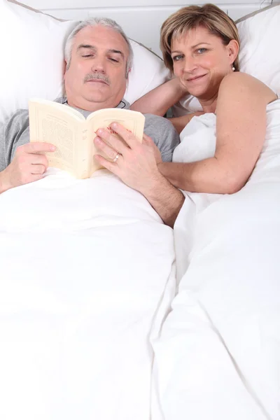 Marido lendo ao lado de sua esposa na cama — Fotografia de Stock