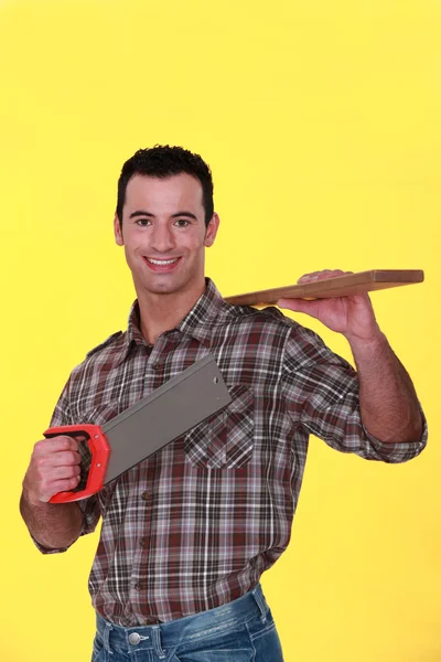 Um carpinteiro carregando uma prancha e uma serra manual . — Fotografia de Stock
