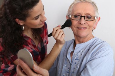 Young woman applying blush on her grandmother's cheeks clipart