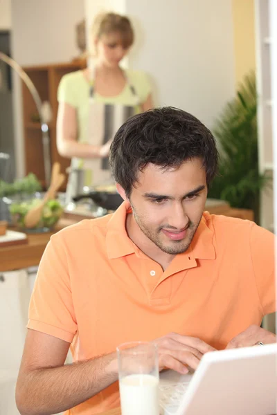 Man op zoek op zijn laptop, terwijl zijn vrouw diner bereidt — Stockfoto