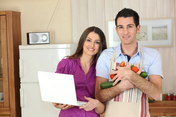 Пара на кухне с ноутбуком и оружием, полным овощей — стоковое фото