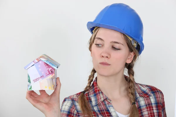 Una trabajadora de la construcción sosteniendo una miniatura de una casa hecha de billetes . — Foto de Stock