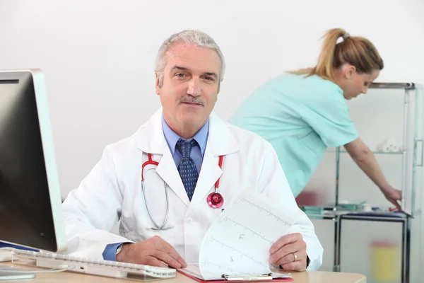 Vedoucí lékař v ordinaci s ošetřovatelkou v pozadí — Stock fotografie