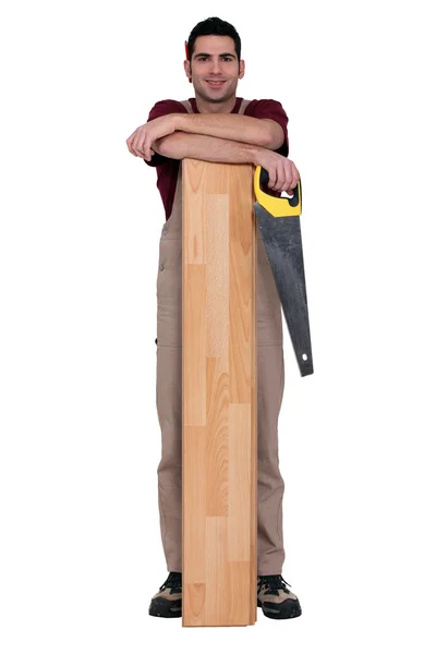 Εργαζόμενος κρατώντας ένα πριόνι και κλίση ενάντια σε μια ξύλινη σανίδα — Φωτογραφία Αρχείου