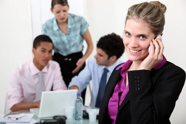 Leende kvinna tar ett samtal framför upptagen kollegor — Stockfoto
