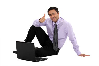 Businessman sitting on the floor with his laptop and making a thumbs up sig clipart