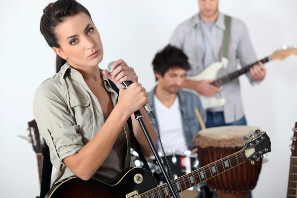 Brunette singer with guitar and microphone and male musicians in background — Stock Photo, Image