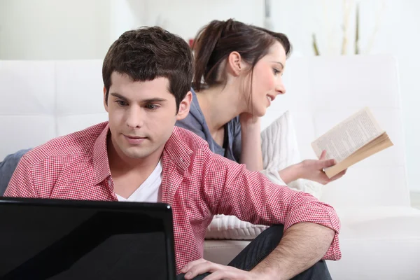 Pareja joven trabajando en el ordenador portátil con la novia leyendo en segundo plano — Foto de Stock