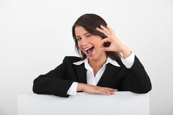 Cheeky young woman in a suit giving the OK sign with a blank board ready fo — Stock Photo, Image