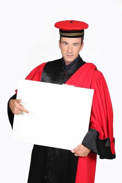 Man in red and black robe and cap pointing at board blank for text — Stock Photo, Image