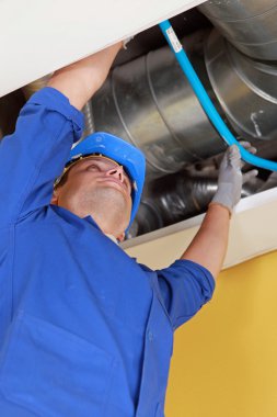 Plumber holding a blue flexible pipe under some air ducts clipart