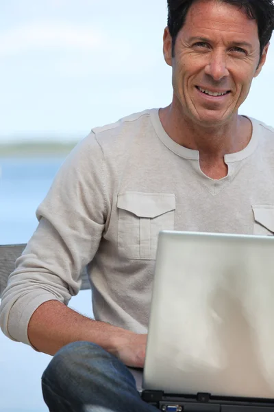 Man with laptop computer sat smiling by water front — Stock Photo, Image