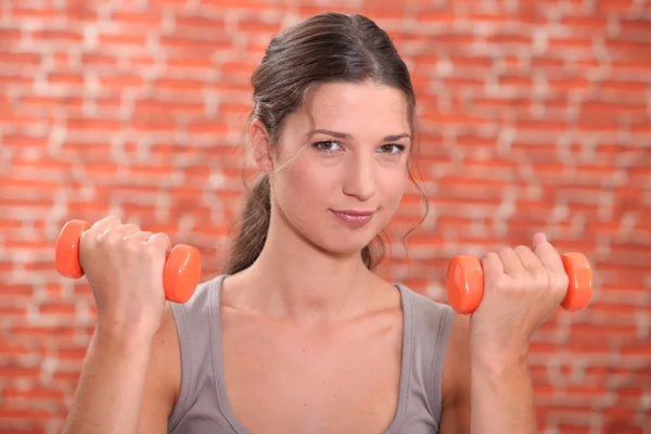 Young woman using hand weights in front of a red brick wall — Stockfoto