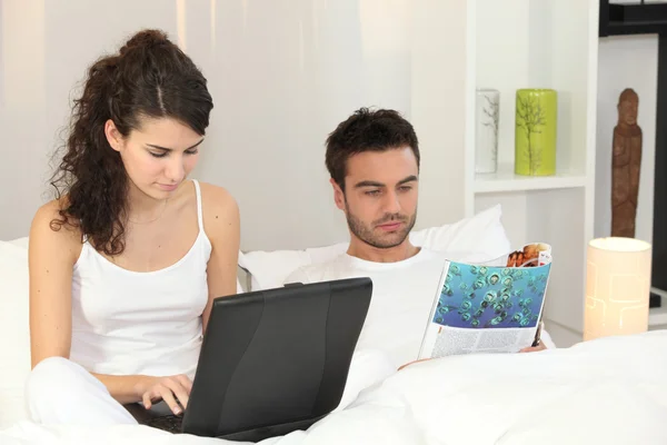 Casal relaxante na cama com revista e laptop — Fotografia de Stock