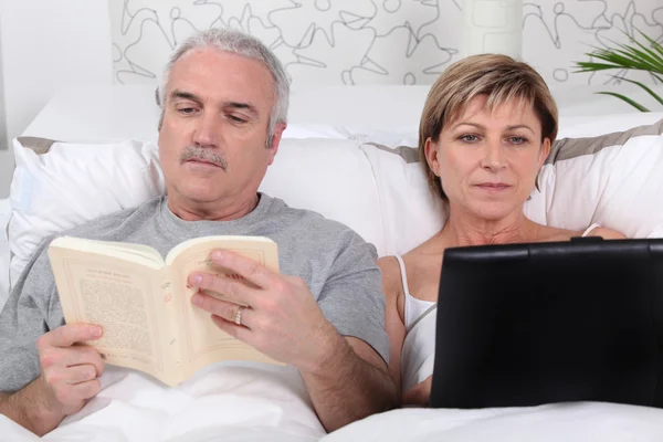 而他的妻子上网冲浪的床上看书的人 — 图库照片
