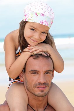 Little girl sitting on father's shoulders on the beach clipart