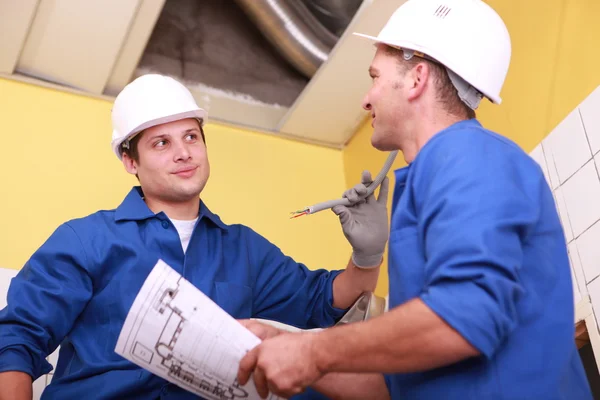 Two workers dressed in blue jumpsuits in a house under construction, one of — Stock Photo, Image